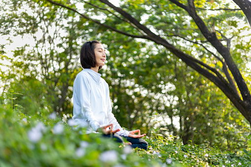 Asian woman is relaxingly practicing meditation yoga in forest full of wild flower in summer to attain happiness from inner peace wisdom with morning light for healthy mind and soul concept