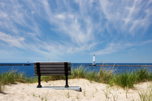A park bench sits on the bay with Frankfort, Michigan lighthouse and a channel marker in the background. Frankfort is located along Michigan's Scenic Route M-22.