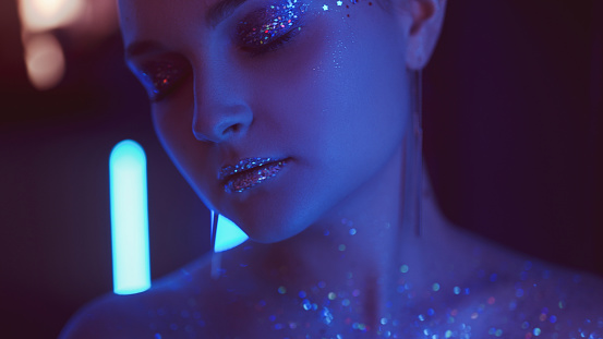 Neon face. Glitter skin. Night party look. Blue purple color light portrait of relaxed woman with closed eyes artistic makeup shimmering eyeshadow lips on dark.