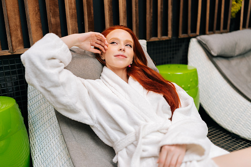 Portrait of satisfied charming young woman in white bathrobe relaxing lying on deck chair after beauty treatment at spa salon, looking away. Pretty redhead female enjoying wellness weekend alone