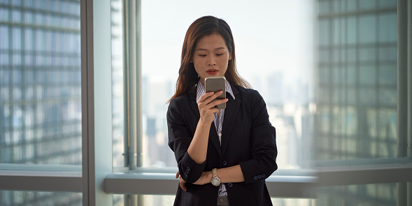 young asian business woman standing by the window looking at cellphone in modern office