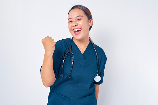 Excited young Asian female professional nurse working wearing a blue uniform standing confident while saying yes and celebrating victory isolated on white background. Healthcare medicine concept