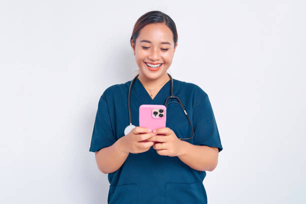 Smiling friendly young Asian woman nurse working wearing a blue uniform using a mobile phone on messaging or using an app isolated on white background. Healthcare medicine concept