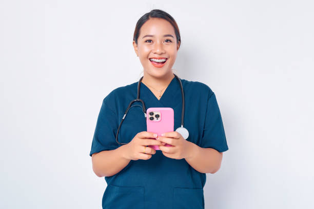 istock Smiling friendly young Asian woman nurse working wearing a blue uniform using a mobile phone on messaging or using an app isolated on white background. Healthcare medicine concept 1482976501