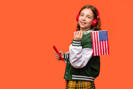 Teen school girl in casual clothes and headphones holds small American flag, or the U.S. flag and smartphone. Languages courses online. Learning American English, student exchange and travel concept