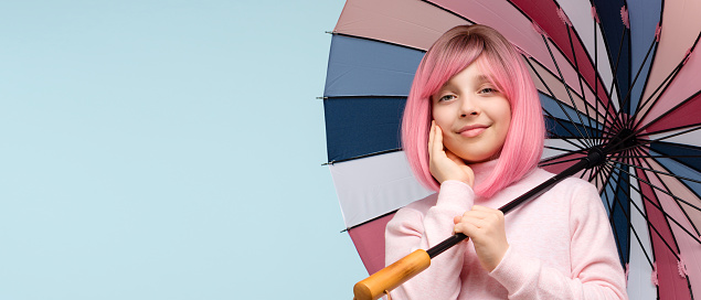 Teen girl with pink hair wig and multicolored umbrella, wears pastel-colored clothes. Cute portrait teenager looking at camera on blue background studio. Banner