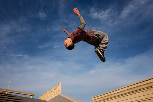 Man doing back somersault against blue sky. Parkour athlete training in city in sportive clothes. Free runner jumping have workout. Active lifestyle. Extreme sports, outdoor freerun