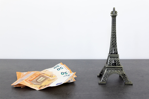 Metal Eiffel Tower statue and fan of 50 euro bills on black table surface against white wall background, conveying a tone of luxury and wealth associated with visiting Paris.