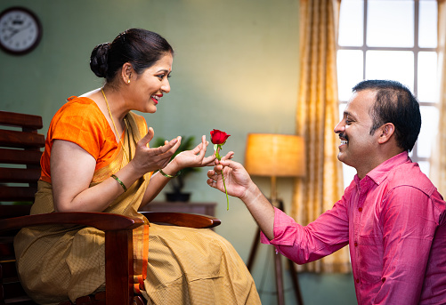 Happy middle aged man proposing his wife by giving red rose at home - concept of wedding anniversary, affection and love emotions