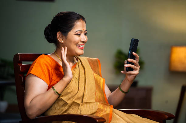 istock Happy indian middle aged woman busy making video calling on mobile phone while sitting on chair at home - concept of technology, relaxation and connection 1482987405