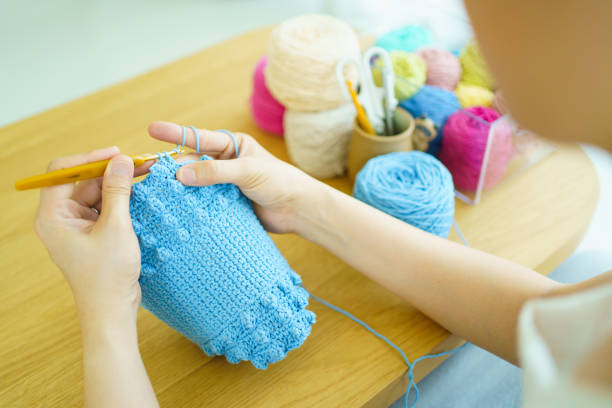 Happy cheerful Asian woman doing a crochet in living room in free time. Skillful Asian craftswoman knitting a handcraft crochet sweater and hat in cozy living room.