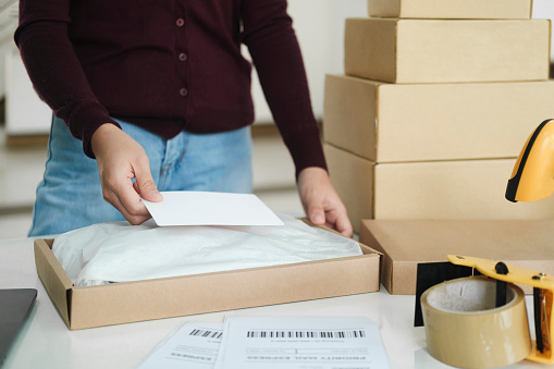 Close up of young female online business owner standing at table with cardboard boxes and laptop at workplace packing packages to online orders preparing for shipment and delivery to customers. Online business, shipment, delivery concept.