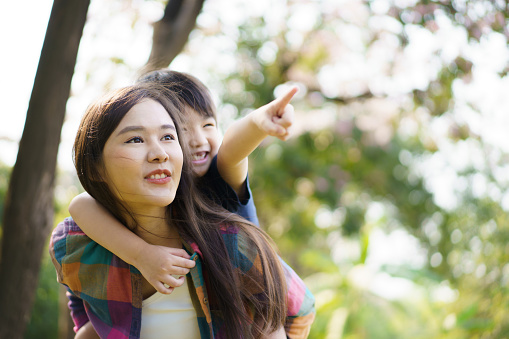 Asian woman and boy playing and walking around the park together with beautiful bokeh and lighting background copyspace. Little cute boy hogging his mother from back and kissing.