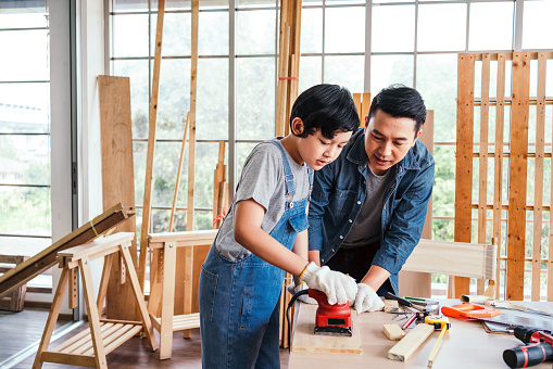 Asian father and son carpentry sanding timber plank to smooth with an electric sander machine together carefully at the home carpentry studio. Carpentry working at a home workshop studio.
