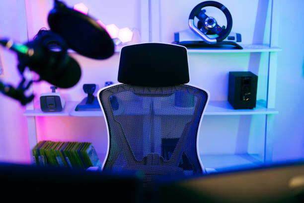 Darkroom for a professional gamer with a comfortable empty gamer chair, computer table, neon lights, keyboard, monitor, controllers and microphone for recording podcasts and live streaming at home.