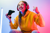 A young Caucasian beautiful woman in a yellow hoodie playing a video game using a joystick and headphones with fun, focus, surprise, and a winning victory face with blue and red neon light.