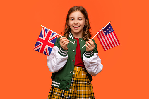 Girl 10-13 y.o. in school outfit on orange background looking at camera. Teen holding UK flag and flag of America. Concept of learning American English and British English.