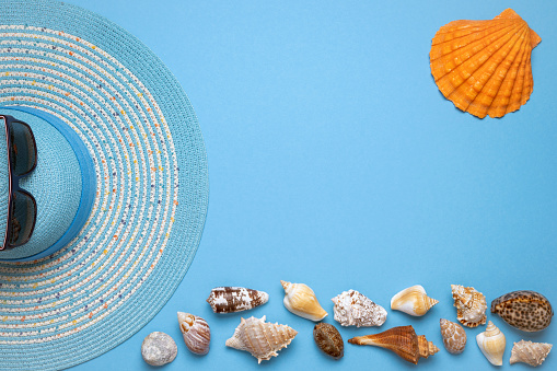 Summer and vacation flat lay with a beauty blue striped woman straw hat, sun glasses, a scallop shell and various seashells at the lower edge of the picture on blue background.