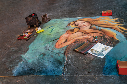 Florence, Italy - August 13, 2016: Pavement artists painting with chalk and pastels on the street of Florence, the Birth of Venus’, masterpiece of the Renaissance,  original painting by Sandro Botticelli 1484.  \nThe original painting depicts the birth of the Roman goddess Venus, who emerges from the sea as a fully grown woman. The painting is considered to be one of the most famous and iconic works of art from the Renaissance period.\nThe painting is set on a beach, with the waves crashing in the background. Venus is depicted standing in a seashell, with her long golden hair cascading down her back. She is surrounded by several figures, including the wind god Zephyrus and his wife Chloris, who is holding a garment to cover Venus. Cupid is also present, aiming his arrow at Venus to signify love and desire.\nBotticelli's use of color and composition is striking in the painting. The soft, pastel colors of the figures and landscape give the painting a dreamlike quality, while the bold outlines and precise details add a sense of realism. The figures are arranged in a graceful and harmonious way, creating a sense of balance and beauty.\nThe Birth of Venus is considered to be a masterpiece of Renaissance art, reflecting the era's fascination with classical mythology, beauty, and nature. The painting has inspired countless artists throughout the centuries and remains a beloved work of art to this day.