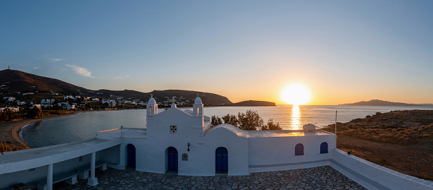 Tinos island, Cyclades Greece. Aerial drone view of Agios Sostis whitewashed orthodox church at sunset, sandy beach, colorful sea, blue sky.