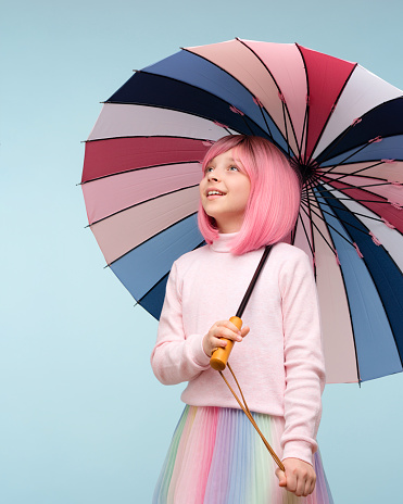 Teen girl with pink hair wig and multicolored umbrella, wears pastel-colored clothes. Teenager surprised, interested looks away on empty space on blue background studio.