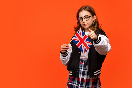 Girl wearing school outfit and holds small UK flag. Teen schoolgirl in round eyeglasses points index finger at camera. Study abroad concept. Learning British English with native speaker.