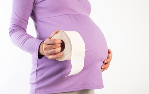 A pregnant girl in a lilac blouse holds toilet paper against the background of her belly. The concept of constipation and diarrhea in pregnant women in connection with the restructuring of the hormonal background. Copy space for text