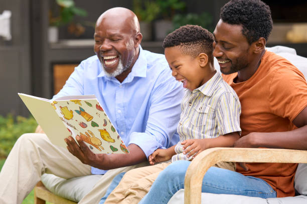 sourire multi-génération famille masculine lisant un livre dans le jardin ensemble - grandparent senior adult child reading photos et images de collection