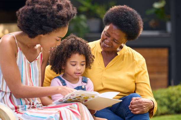 livre de famille féminine multigénérationnelle souriante lisant ensemble dans le jardin - grandparent senior adult child reading photos et images de collection