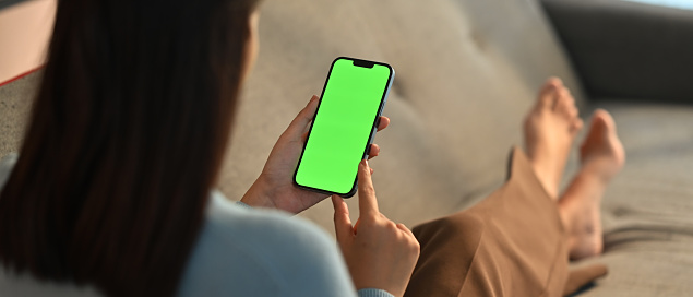 Young woman home use green screen mockup Smartphone, her sitting on sofa in living room