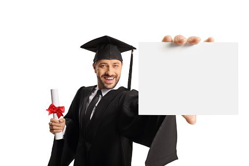 Smiling male student in a graduation gown holding a diploma and showing a blank card isolated on white background