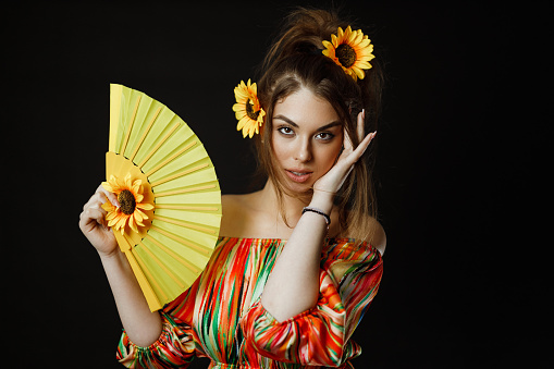 Portrait of a beautiful young smiling woman with sunflowers in her hair holding a yellow color fan, studio photoshoot in front of a black background