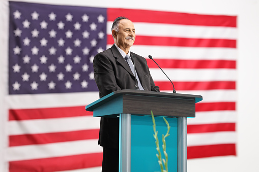 Low angle view at the serious young American politician looking away during his speech at the debates, standing behind the pedestal on a blue background with two American flags