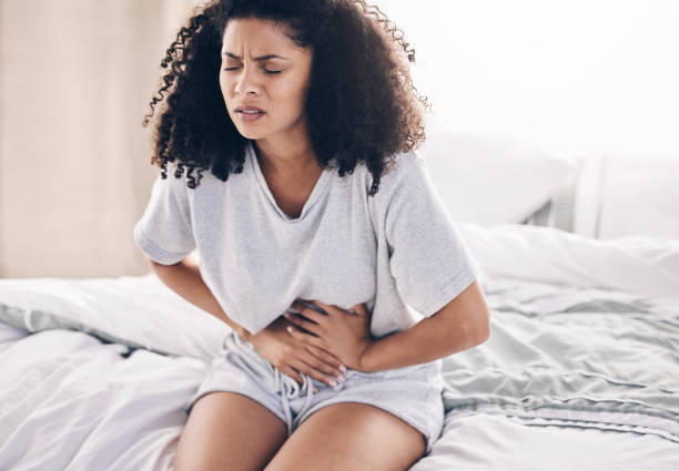 Douleurs abdominales, règles et femme noire au lit avec crampes abdominales, menstruations et maux d’estomac. Santé, urgence médicale et fille avec problème de digestion, crise du syndrome prémenstruel et endométriose dans la chambre - Photo