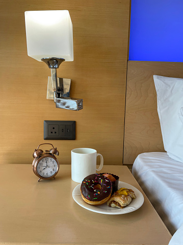 Stock photo showing close-up view of retro, double bell, analogue alarm clock with black numerals, hour and minute hands on beechwood bedside table with white coffee mug and white plate containing a chocolate glazed doughnut, a muffin in paper cake case and a Danish pastry. Home furnishings and bedroom interior design.