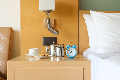 Stock photo showing close-up view of retro, double bell, analogue alarm clock with black numerals, hour and minute hands on beechwood bedside table with white coffee cup on saucer, stainless steel coffee and milk pots and white plate containing cookies. Home furnishings and bedroom interior design.