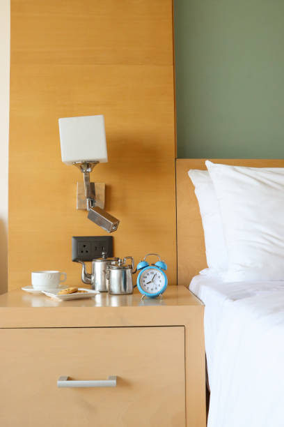 istock Image of nightstand blue metal case, retro alarm clock, double bell, analogue display clock, white coffee cup and saucer, plate of biscuits, stainless steel coffee and milk pots, double bed with white bed sheets and pillows, electric wall lamp 1483021939