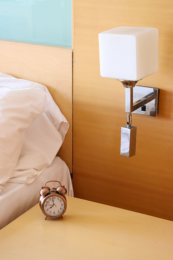 Stock photo showing close-up view of electric wall lamp over retro, double bell, analogue alarm clock with black numerals, hour and minute hands on beechwood bedside table. Home furnishings and bedroom interior design.
