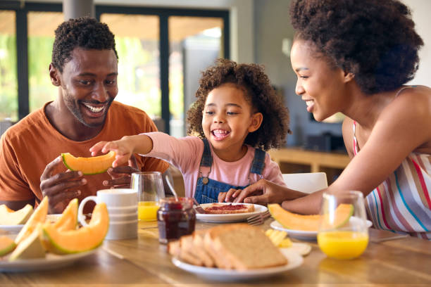 Família filmada com pais e filha em casa tomando café da manhã espalhando geleia no pão na mesa - foto de acervo