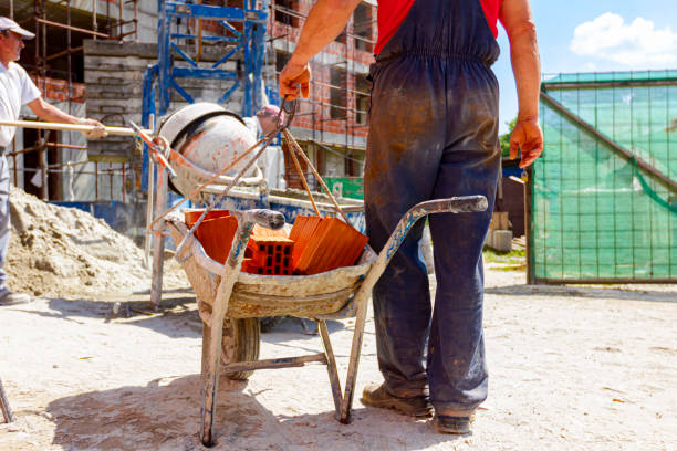 Construction worker is attaching crane hooks to industrial tipping barrow full of red bricks in the background is mortar mixer machine and edifice under construction with scaffold.