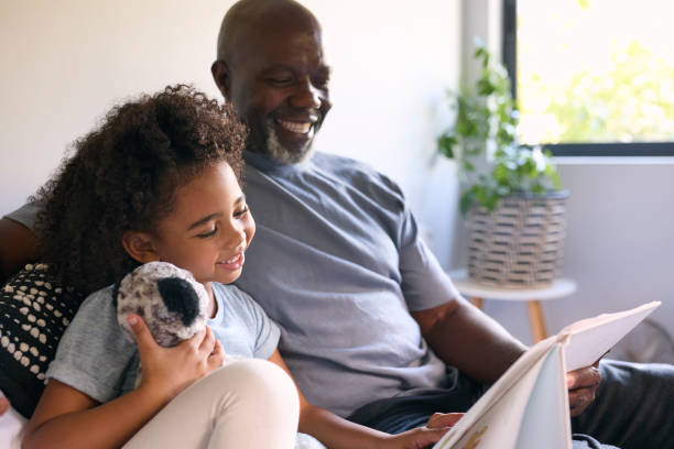 istock Grandfather Sitting On Sofa At Home Reading Book With Granddaughter 1483025708