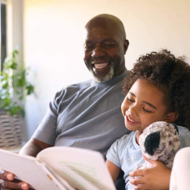 istock Grandfather Sitting On Sofa At Home Reading Book With Granddaughter 1483026445