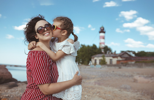 little girl of 5 years kisses happy mother on beach next to lighthouse in summer