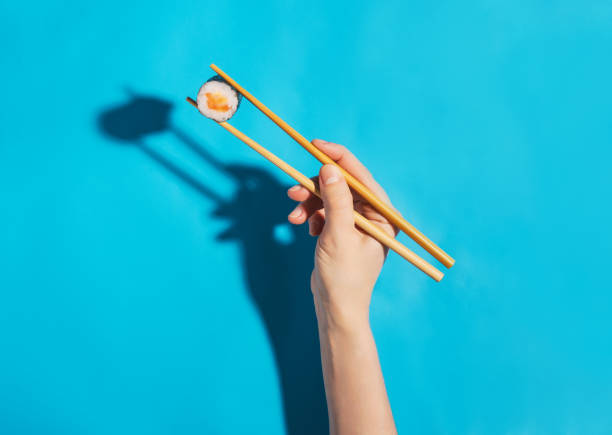 hand with chopsticks holding salmon maki sushi roll on a blue background - food sushi seafood maki sushi imagens e fotografias de stock