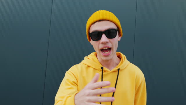 Young man caucasian Singing song moves gesturing with his hands to camera close-up smiling blogger recording video content beanie wearing yellow hoodie grey wall in summer. Younger generation. Life