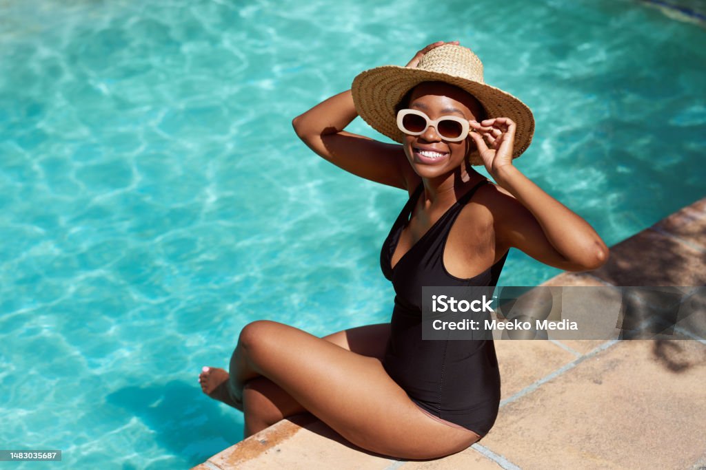 Portrait of young Black woman sitting with legs in pool, holding sunhat posing Portrait of young Black woman sitting with legs in pool, holding sunhat posing. High quality photo Swimming Pool Stock Photo