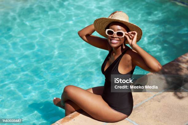 Portrait Of Young Black Woman Sitting With Legs In Pool Holding Sunhat Posing Stock Photo - Download Image Now