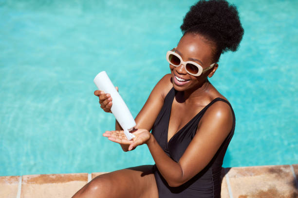 Beautiful Black woman applies sunscreen sitting by pool in summer sun stock photo
