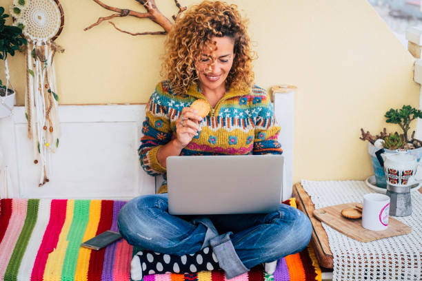 istock Cheerful young adult woman using computer sitting and comfy eating cookie. Happy home lifestyle and surfing the net. Modern people work on laptop in leisure online activity. Alternative office 1483036755