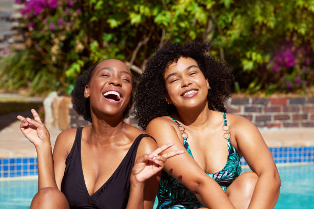 Two Black friends laugh in the sunshine sitting at the pool, summer staycation